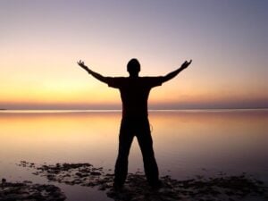 A person standing with arms outstretched in front of a serene ocean and sunset